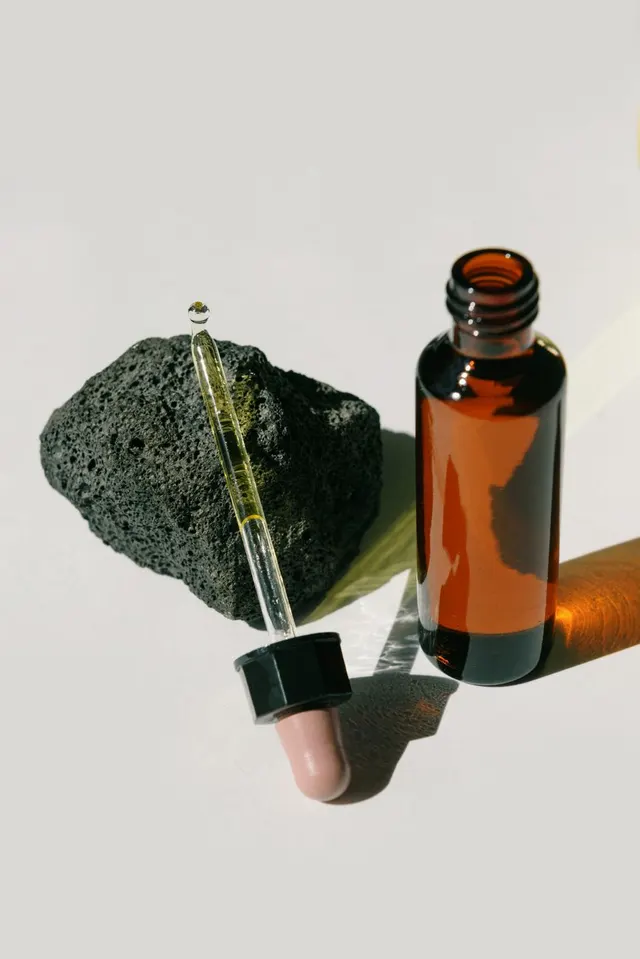 Frasco de cristal ámbar abierto junto a una roca volcánica negra y un cuentagotas de vidrio con una gota de aceite en la punta