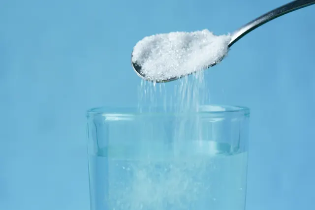Una cuchara metálica vertiendo azúcar blanco en un vaso de agua sobre un fondo azul