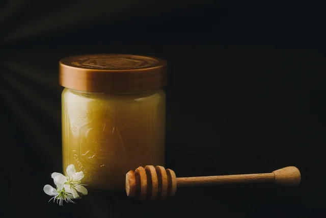 Tarro de cristal de miel cremosa con tapa marrón y un cazo de madera para miel al lado