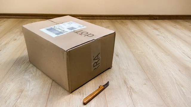 Una caja de cartón cerrada con una etiqueta de envío, en el suelo de madera junto a un cúter