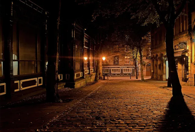 Calle adoquinada de noche iluminada por farolas de luz anaranjada, con árboles y fachadas de piedra